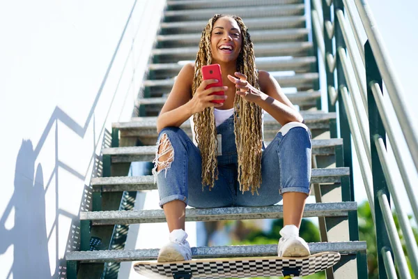 Zwart meisje met gekleurde vlechten, zittend met een smartphone en een skateboard. — Stockfoto
