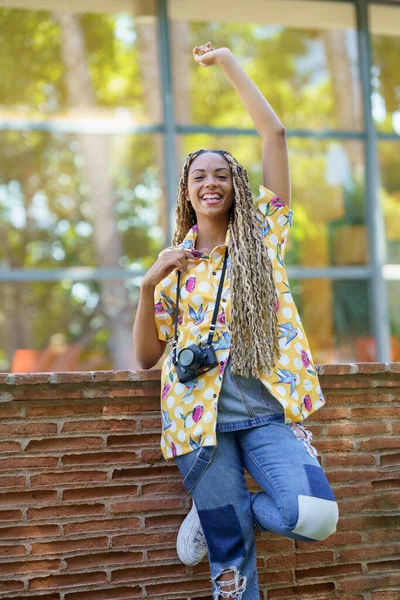 Zwarte vrouw met Afrikaanse vlechten, die haar arm optilt van vreugde. Meisje met een camera. — Stockfoto
