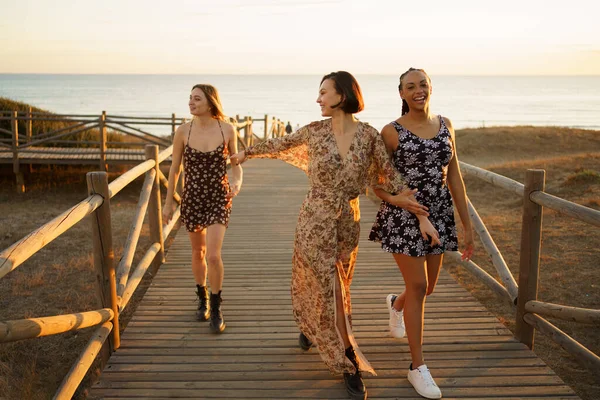 Vrolijke gevarieerde vriendinnen wandelen op voetgangersbrug in de buurt van zee — Stockfoto