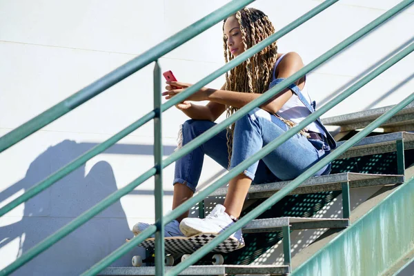 Zwarte vrouw met gekleurde vlechten, raadpleegt haar smartphone met haar voeten rustend op een skateboard. — Stockfoto