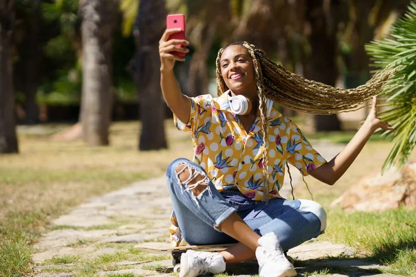 Donna nera con i capelli intrecciati scattare un selfie con uno smartphone seduto sul suo skateboard — Foto Stock