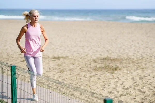 Пожилая женщина занимается спортом, чтобы поддерживать форму. Зрелая женщина бежит вдоль берега пляжа. — стоковое фото