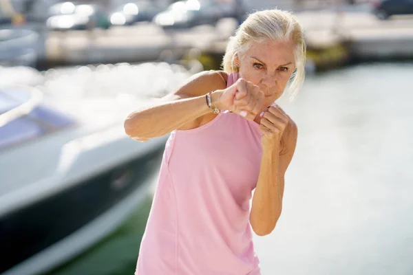 Зрелая женщина занимается теневым боксом на открытом воздухе. Старшая женщина занимается спортом в прибрежном порту — стоковое фото