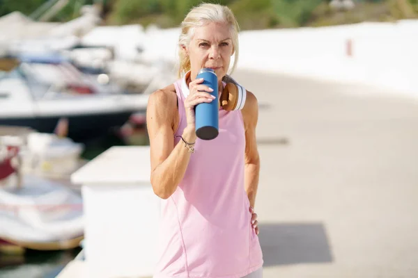 Зрелая спортсменка в спортивной одежде пьет воду из металлической фитнес-бутылки на открытом воздухе. — стоковое фото