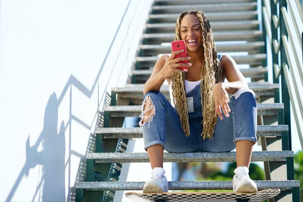 Zwarte vrouw met gekleurde vlechten, raadpleegt haar smartphone met haar voeten rustend op een skateboard. — Stockfoto