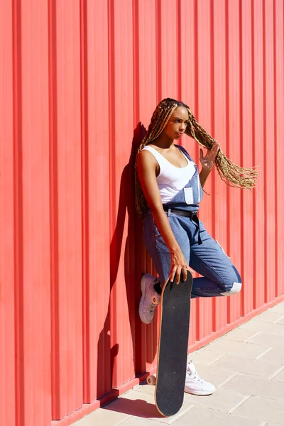 Mujer negra vestida casual, con un monopatín sobre fondo rojo de pared urbana. — Foto de Stock