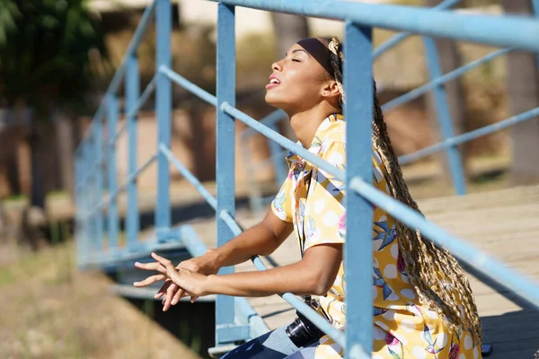 Saçları örülü siyah bir kız. Köprüde oturmuş, gözleri kapalı güneşin tadını çıkarıyor.. — Stok fotoğraf