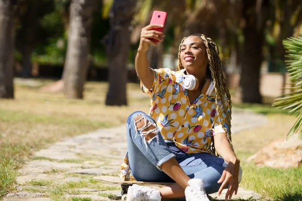 Donna africana con i capelli intrecciati che si fa un selfie con uno smartphone seduto sul suo skateboard — Foto Stock