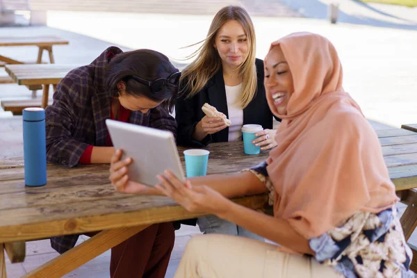 Contenidos jóvenes amigas multirraciales riendo mientras comparten la tableta en la cafetería —  Fotos de Stock