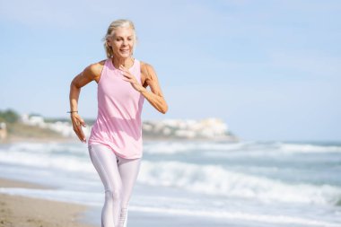 Yaşlı kadın formda kalmak için spor yapıyor. Kumsalda koşan olgun bir kadın..