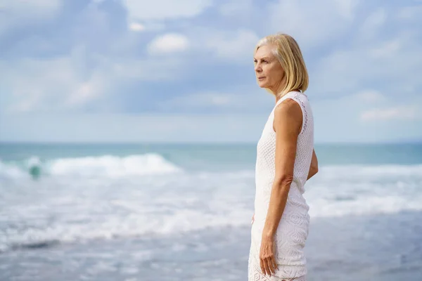 Volwassen vrouw die sereen naar de zee staart. Oudere vrouw die op een locatie aan zee staat — Stockfoto