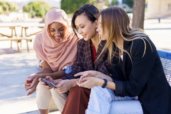 Positive multiethnische Freundinnen teilen Smartphone, während sie sich auf Bank im Park ausruhen — Stockfoto