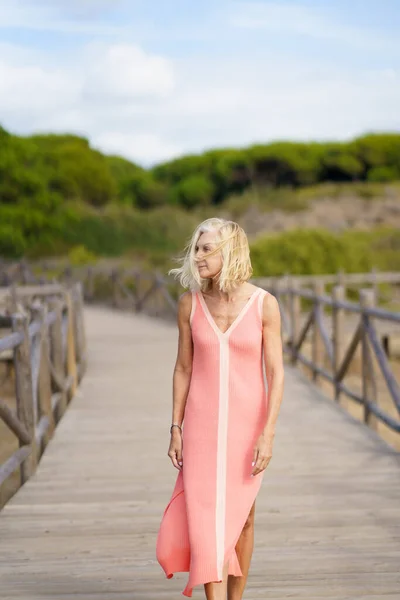 Mulher madura bonita andando ao longo de um caminho de madeira perto da praia., vestindo um vestido laranja agradável. — Fotografia de Stock