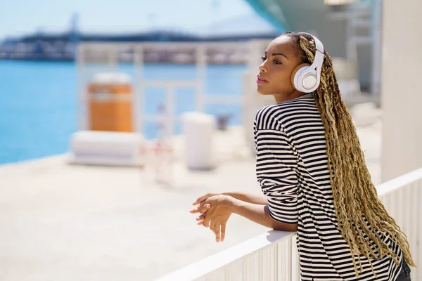 Jonge zwarte vrouw die naar muziek luistert terwijl ze geniet van het uitzicht op de zeehaven. — Stockfoto