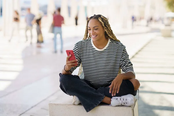 Zwarte vrouw met behulp van een smartphone zitten op een bank buiten, het dragen van haar haar in vlechten. — Stockfoto