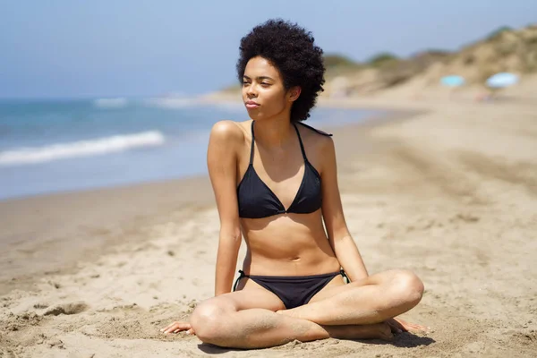 Ontspannen zwarte vrouw in bikini zittend op het zand van een tropisch strand uitkijkend op zee. — Stockfoto