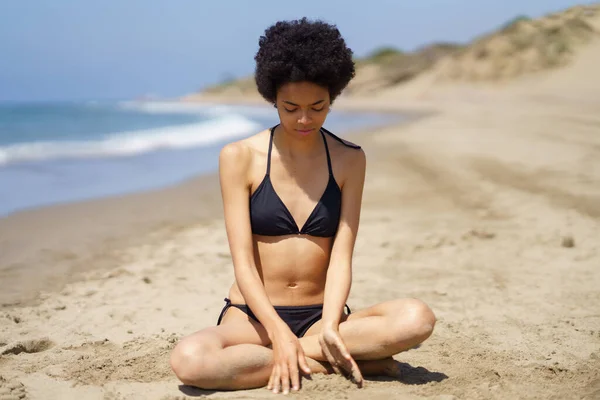 Позитивная черная девушка в бикини, сидящая на песке пляжа, задумчиво глядя на песок. — стоковое фото