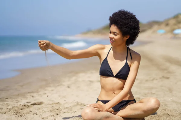 Positive black woman in bikini sitting playing with sand on the beach. — Stock Photo, Image