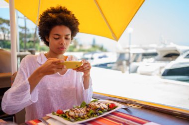 Genç siyah bir kadın, afro saçlı, arka planda tekneleri olan bir sahil bölgesinde bir barda akıllı telefonuyla keçi peyniri salatası tabağını fotoğraflıyor..
