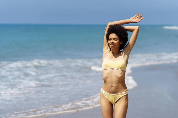 幸せなアフリカ系アメリカ人女性は太陽の下で彼女の休日を楽しむためにビーチで腕を上げています 若いです黒女性身に着けている黄色ビキニ — ストック写真