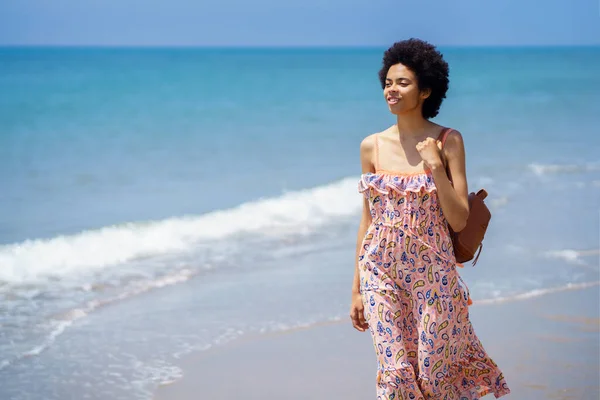 Afro-haired black woman in summer dress, walking on the sand of a tropical beach. Girl enjoying her holiday in a coastal area.