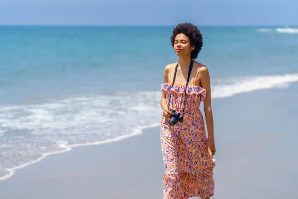 Positive black woman walking on the sand of a tropical beach in a summer dress and slr camera. Girl enjoying her holiday in a coastal area.