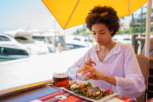 Genç siyahi bir kadın, afro saçlı, arka planda tekneleri olan, kıyı bölgesindeki bir barda keçi peynirli salatasına tuz döküyor..