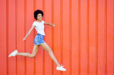 Kırmızı bir şehir duvarına karşı kollarını kaldırarak atlarken kameraya bakan mutlu siyah kadın. Tam vücut fotoğrafı.