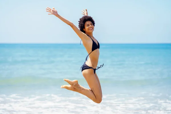 陽気なアフリカ系アメリカ人女性 旅行者でビキニを見ますカメラ ジャンプしながら腕を持ち上げ反対日当たりの良い日に海 — ストック写真