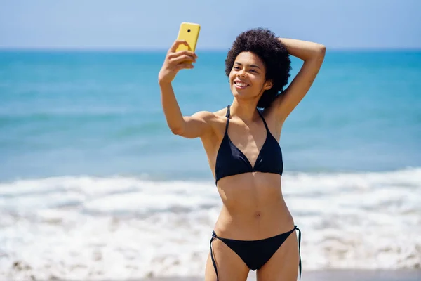 楽観的なフィットアフリカ系アメリカ人女性旅行者で水着姿で携帯電話で自分の肖像画を取ります頭の後ろに手で夏に波の近くに立って海 — ストック写真