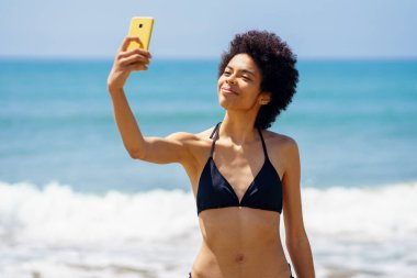 İyimser Afrikalı Amerikalı kadın turist yaz tatilinde deniz kenarında dururken cep telefonuyla kendi portresini çekiyor.