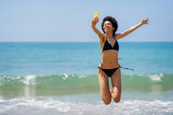 熱帯リゾートの海の近くのスマートフォンに自分の肖像画を取りながら 腕を上げてジャンプ水着で楽しいアフリカ系アメリカ人女性観光客 — ストック写真