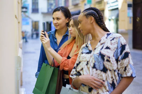 ショッピングバッグを持つ多民族女性の友人のサイドビューは 街の店の近くに立っている間 スマートフォンでショーケースの写真を撮る — ストック写真