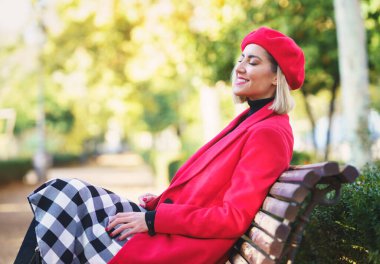 Kırmızı ceketli, bereli, gülen bir kadın, ahşap bir bankta oturuyor sonbahar parkında gözleri kapalı bulanık arka planda.