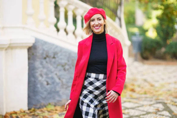 Happy female in red outfit and beret looking at camera while strolling on path near fence on street with trees in city