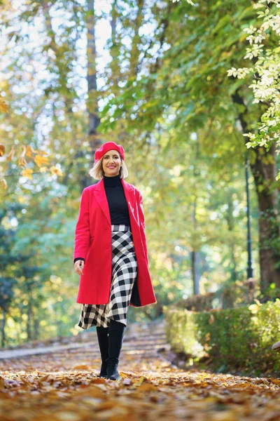 Full body of positive female in red coat and beret looking at camera while strolling on path with fallen leaves on autumn day