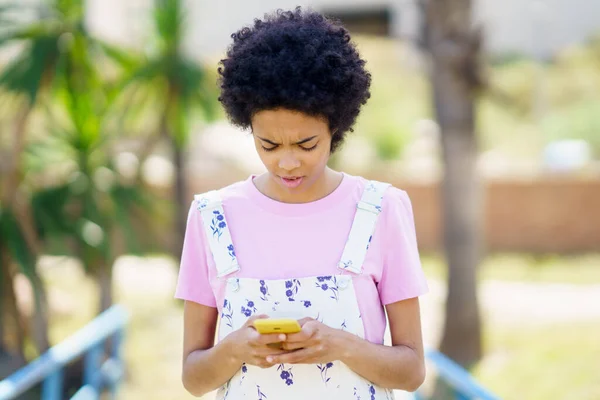 Puzzled African American female texting message on cellphone while standing on street with trees against blurred background on summer day