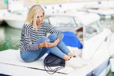 Deniz limanında oturan akıllı telefonunu kullanan olgun bir kadın. Yaşlı bir kadın deniz kenarı inzivasında emekliliğinin tadını çıkarıyor..