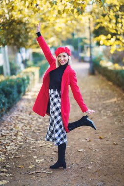 Full body of positive female in classic outfit with bright beret looking at camera with raised arm while standing on one leg in park