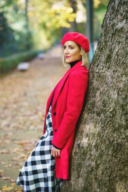 Side view of stylish female wearing red coat, and beret leaning on tree trunk on pathway covered with fallen dry leaves in autumn park and looking away