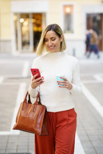 Happy female in stylish outfit and handbag browsing modern cellphone while strolling with cup of drink on street