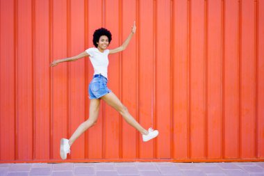 Full body side view of cheerful African American female in denim shorts looking at camera while jumping on red background