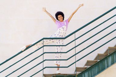 Full body of cheerful African American female, in casual clothes standing on stairway with leg on railing while looking at camera with raised arms against white background