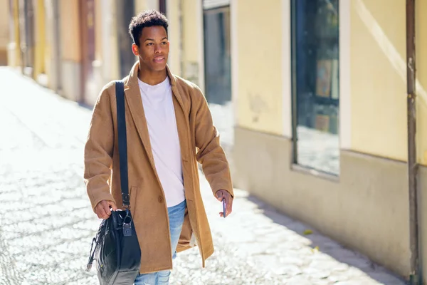 Young black man walking down the street carrying a briefcase and a smartphone. Cuban guy in urban background.