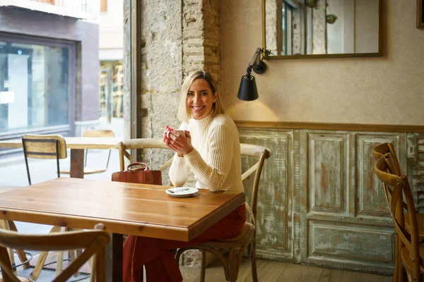 明るいカフェで木製のテーブルに座っている間に笑顔でカメラを見て熱い飲み物のカップを持つ陽気な女性 — ストック写真