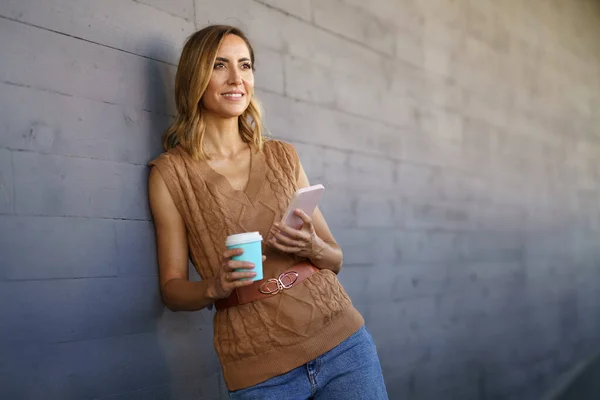 中年妇女在办公室附近喝咖啡休息 白人女性拿着纸杯 手里拿着智能手机 — 图库照片