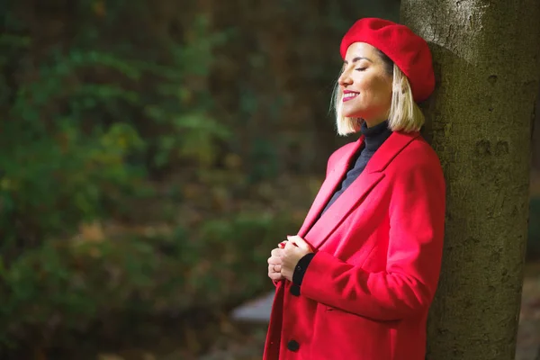 赤いベレー帽をかぶったスタイリッシュな女性と木の幹にもたれかかっているジャケットの側面図と公園の日光から目を閉じる — ストック写真