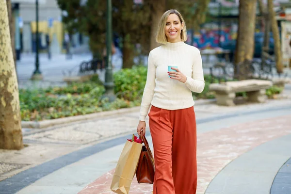 ショッピングバッグやハンドバッグを持ち 公園でテイクアウト飲料のカップを持っているカジュアルな服の正の女性 — ストック写真