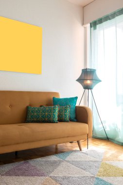 Turkuaz yastıklı ve modern lambalı hardal kanepe detayı. Duvarda bir tablo var. İçeride kimse yok.