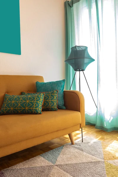 Turkuaz yastıklı ve modern lambalı hardal kanepe detayı. Duvarda bir tablo var. İçeride kimse yok.